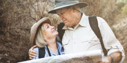 זוג מבוגרים מטיילים עם מפה מחובקים מסתכלים אחד על השנייה שניהם עם כובע ותיק על הגב ברקע של טיול בטבע